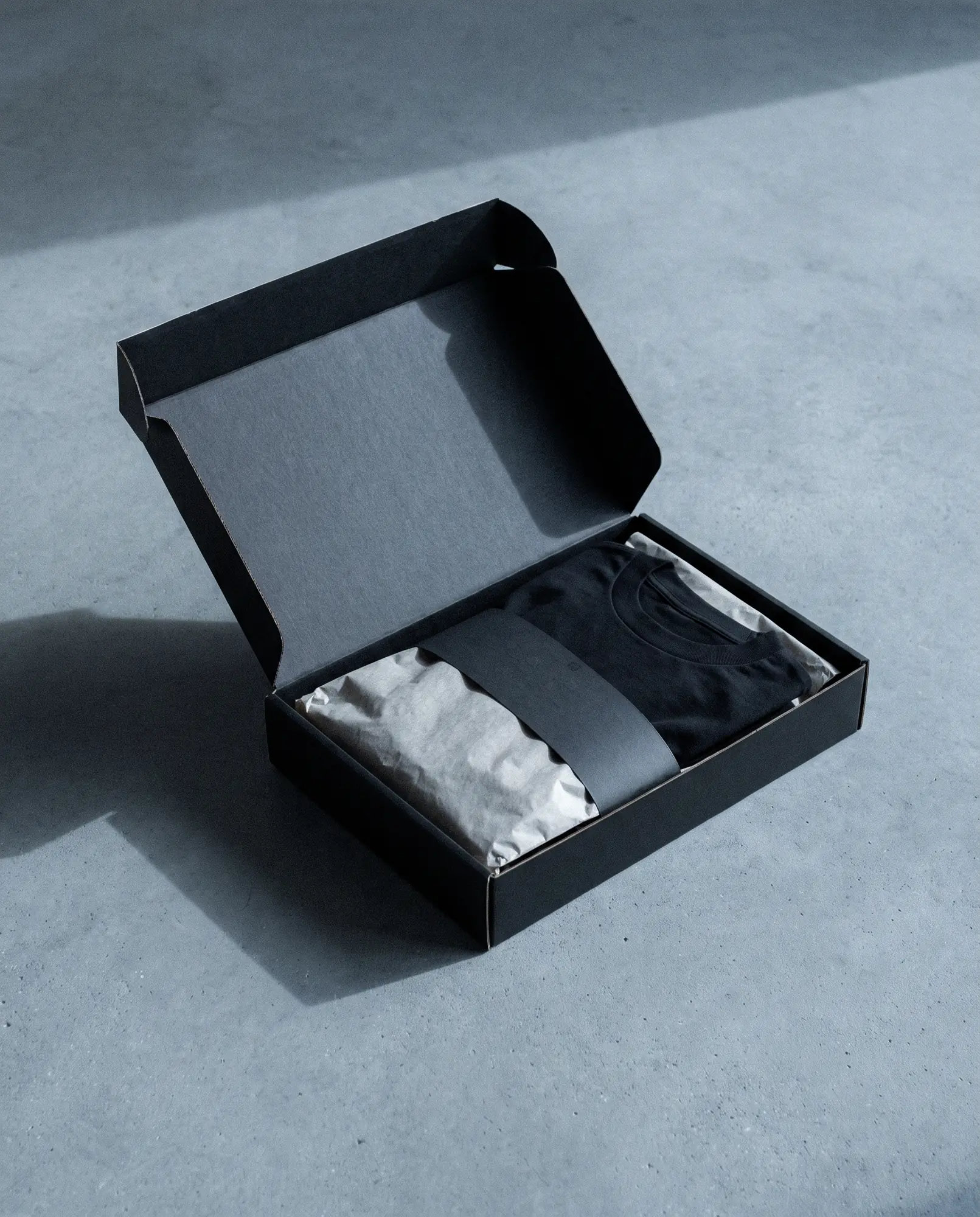 A plain unbranded black mailing bag containing a packed t-shirt, on grey concrete.