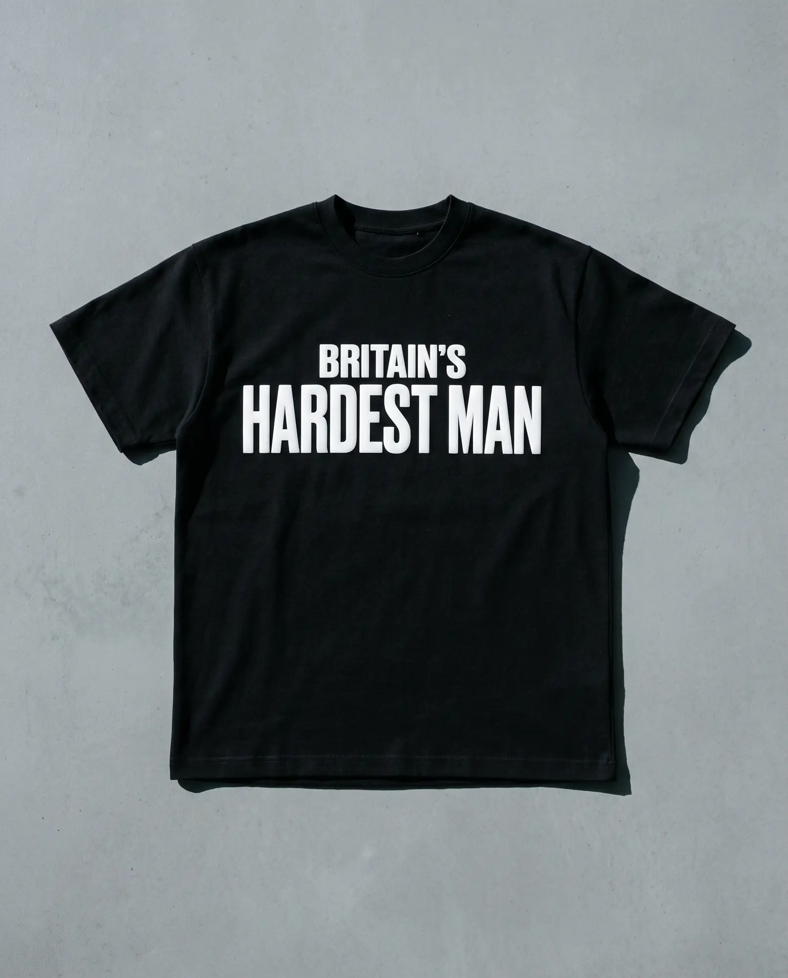 A heavyweight black t-shirt laid flat on grey concrete, BRITAIN'S HARDEST MAN screen printed across the chest in white.