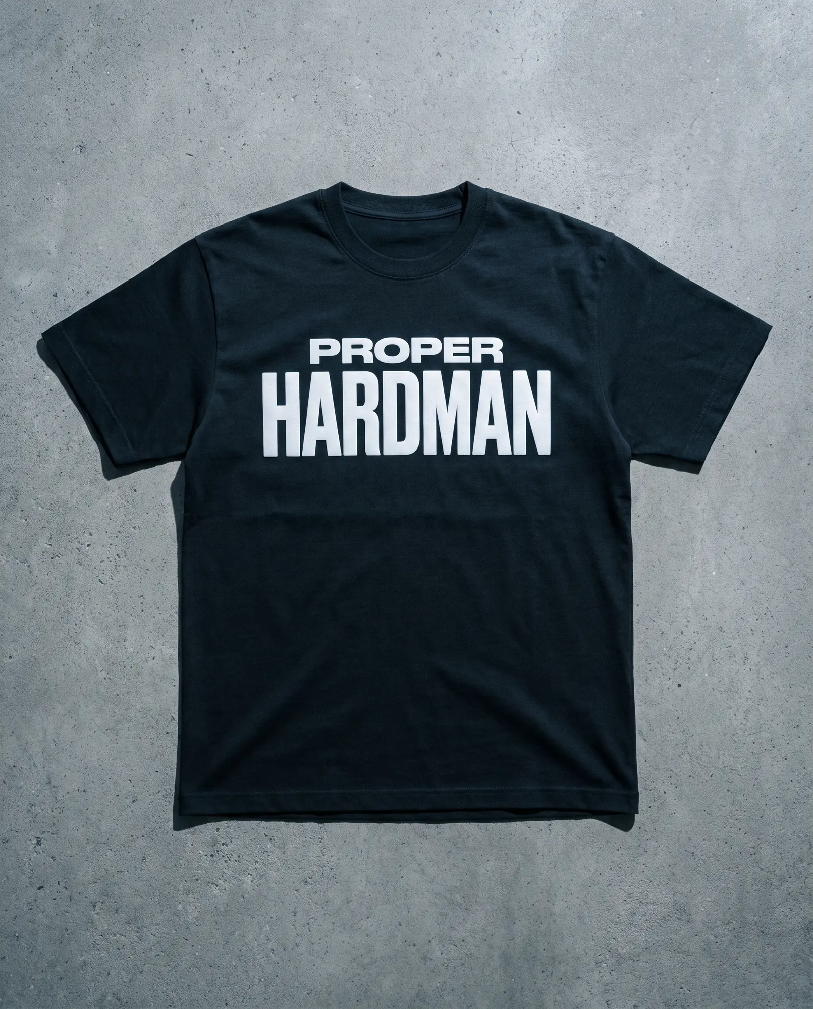 A heavyweight black t-shirt laid flat on grey concrete, PROPER HARDMAN screen printed across the chest in white.