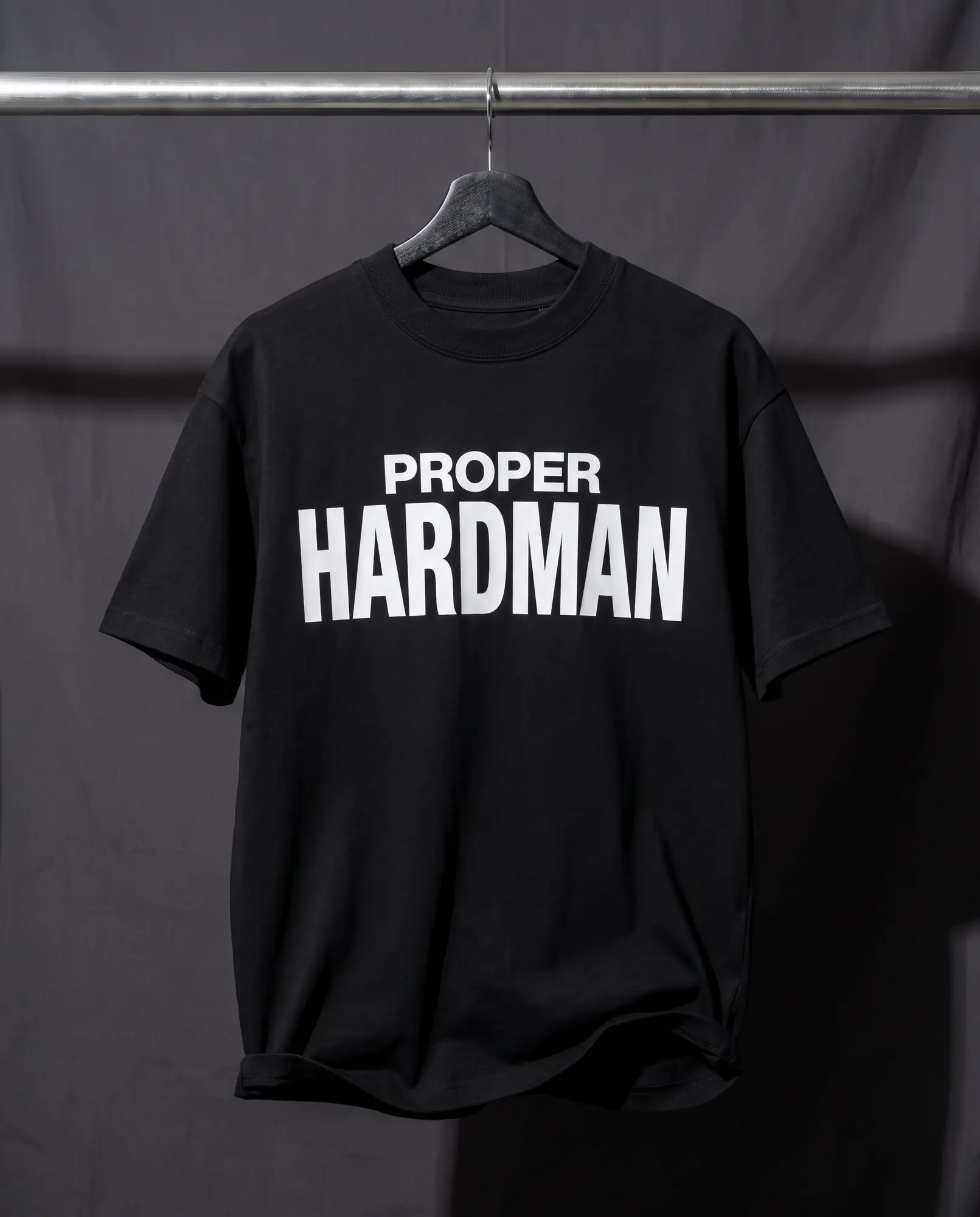 A heavyweight black t-shirt hanging from a brushed steel rail under hard directional light, PROPER HARDMAN screen printed across the chest in white.