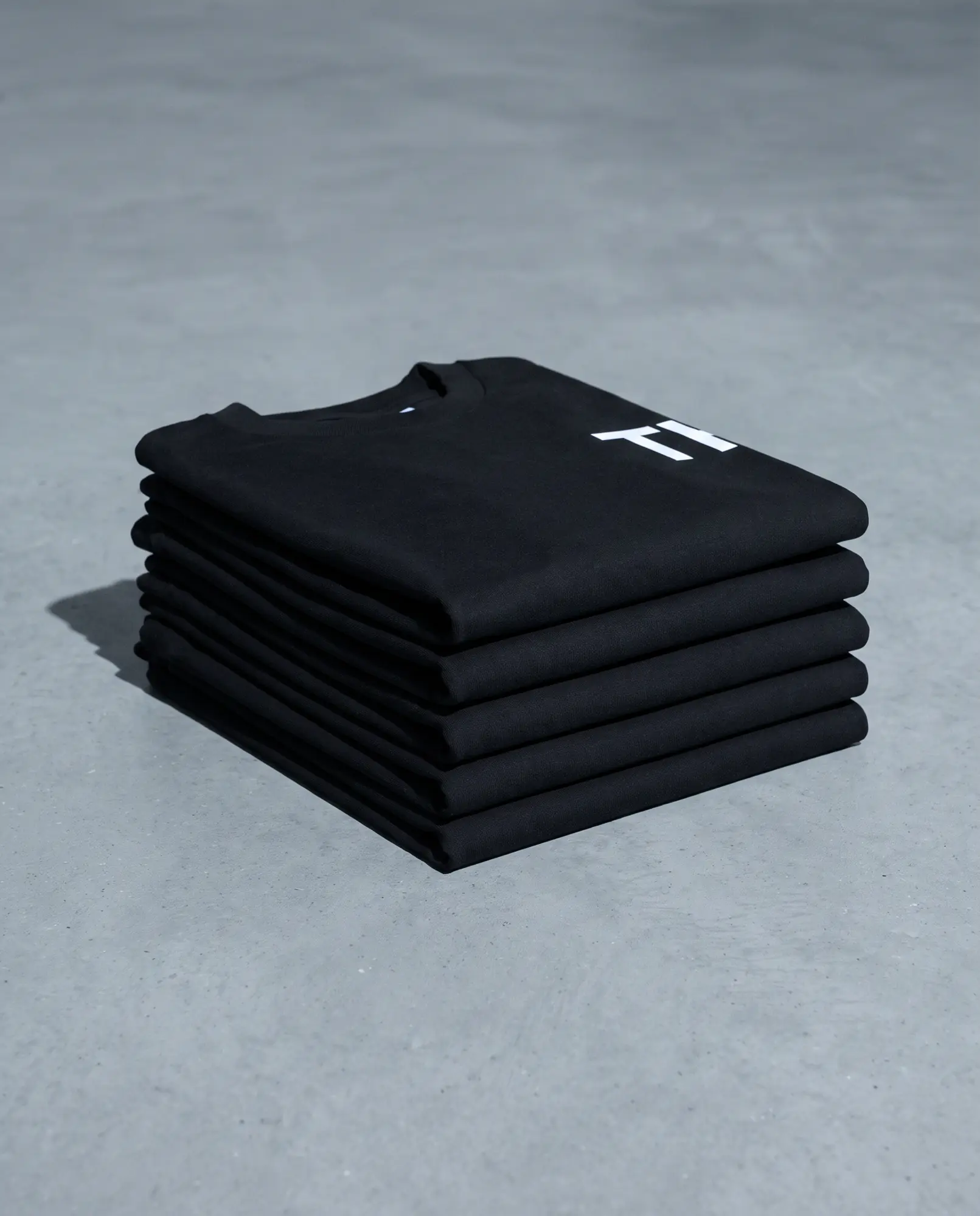 Folded black t-shirts stacked on grey concrete.